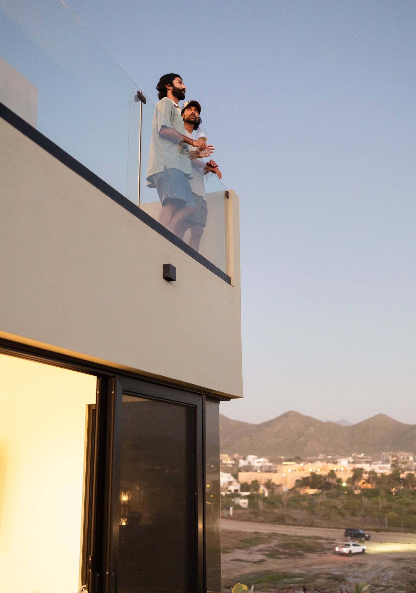 Two people standing at the rooftop rail of the penthouse at dusk, hills and town below